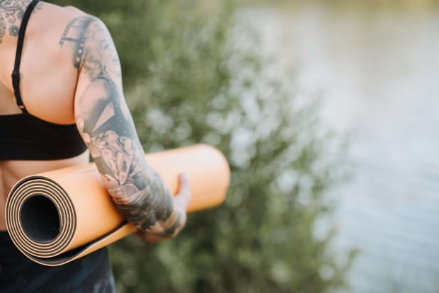 A tattooed woman holding an orange yoga mat under her arm, looking over a body of water.
